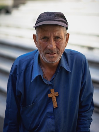 An older man wearing a dark cap and a blue collared shirt stands facing the camera. He has a weathered complexion and a gray beard. Around his neck, he wears a wooden cross pendant. The background is out of focus, consisting of steps or a similar structure.