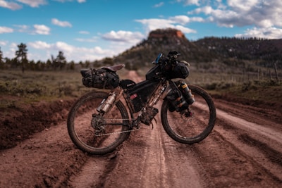 A bicycle equipped with various packs and gear stands on a muddy dirt road, surrounded by a natural landscape with trees and hills in the background. The sky is partly cloudy, suggesting a pleasant weather scene. The bicycle appears to be set up for a long-distance or adventure ride.