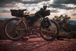 A rugged mountain bike loaded with gear is parked on a rocky trail. The setting sun casts a warm glow, and the sky is filled with dramatic clouds. Trees are visible in the background, contributing to a sense of adventure and exploration.