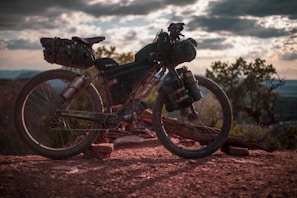 A rugged mountain bike loaded with gear is parked on a rocky trail. The setting sun casts a warm glow, and the sky is filled with dramatic clouds. Trees are visible in the background, contributing to a sense of adventure and exploration.