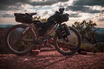 A rugged mountain bike loaded with gear is parked on a rocky trail. The setting sun casts a warm glow, and the sky is filled with dramatic clouds. Trees are visible in the background, contributing to a sense of adventure and exploration.