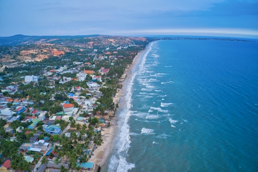 A coastal town stretches along the shoreline with numerous houses and buildings bordered by a vast expanse of ocean. The sea exhibits gentle waves touching the sandy beach, while lush greenery and hills frame the backdrop. The view captures a blend of urban and natural landscapes under a calm blue sky.