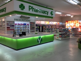 A clean and well-organized pharmacy interior with bright lighting. The central counter is labeled for prescriptions and is surrounded by shelves stocked with various pharmaceutical products. The predominant color scheme involves green and white, reflecting the pharmacy's branding. The aisles are spacious and neatly arranged with products.