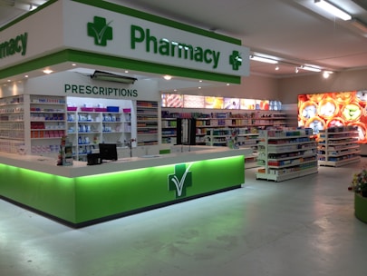 A clean and well-organized pharmacy interior with bright lighting. The central counter is labeled for prescriptions and is surrounded by shelves stocked with various pharmaceutical products. The predominant color scheme involves green and white, reflecting the pharmacy's branding. The aisles are spacious and neatly arranged with products.