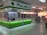 A clean and well-organized pharmacy interior with bright lighting. The central counter is labeled for prescriptions and is surrounded by shelves stocked with various pharmaceutical products. The predominant color scheme involves green and white, reflecting the pharmacy's branding. The aisles are spacious and neatly arranged with products.