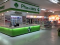 A clean and well-organized pharmacy interior with bright lighting. The central counter is labeled for prescriptions and is surrounded by shelves stocked with various pharmaceutical products. The predominant color scheme involves green and white, reflecting the pharmacy's branding. The aisles are spacious and neatly arranged with products.