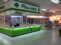 A clean and well-organized pharmacy interior with bright lighting. The central counter is labeled for prescriptions and is surrounded by shelves stocked with various pharmaceutical products. The predominant color scheme involves green and white, reflecting the pharmacy's branding. The aisles are spacious and neatly arranged with products.
