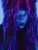 A person is partially covered by a furry garment with neon purple, pink, and blue colors. The lighting creates a dramatic and mysterious atmosphere, with the person's intense gaze directed at the viewer.