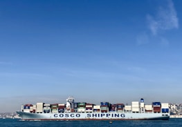 A large cargo ship with the name 'COSCO SHIPPING' is carrying numerous stacked shipping containers on calm blue waters. The background shows a distant coastline under a clear, blue sky.