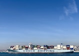 A large cargo ship with the name 'COSCO SHIPPING' is carrying numerous stacked shipping containers on calm blue waters. The background shows a distant coastline under a clear, blue sky.