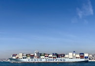 A large cargo ship with the name 'COSCO SHIPPING' is carrying numerous stacked shipping containers on calm blue waters. The background shows a distant coastline under a clear, blue sky.