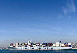 A large cargo ship with the name 'COSCO SHIPPING' is carrying numerous stacked shipping containers on calm blue waters. The background shows a distant coastline under a clear, blue sky.