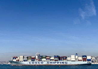 A large cargo ship with the name 'COSCO SHIPPING' is carrying numerous stacked shipping containers on calm blue waters. The background shows a distant coastline under a clear, blue sky.
