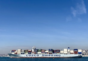 A large cargo ship with the name 'COSCO SHIPPING' is carrying numerous stacked shipping containers on calm blue waters. The background shows a distant coastline under a clear, blue sky.