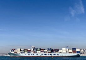A large cargo ship with the name 'COSCO SHIPPING' is carrying numerous stacked shipping containers on calm blue waters. The background shows a distant coastline under a clear, blue sky.