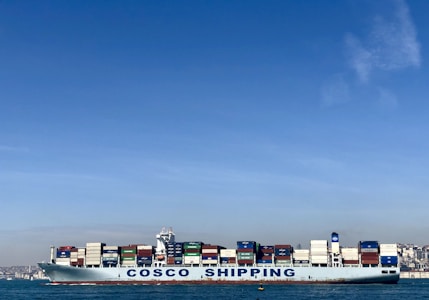 A large cargo ship with the name 'COSCO SHIPPING' is carrying numerous stacked shipping containers on calm blue waters. The background shows a distant coastline under a clear, blue sky.