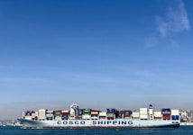 A large cargo ship with the name 'COSCO SHIPPING' is carrying numerous stacked shipping containers on calm blue waters. The background shows a distant coastline under a clear, blue sky.
