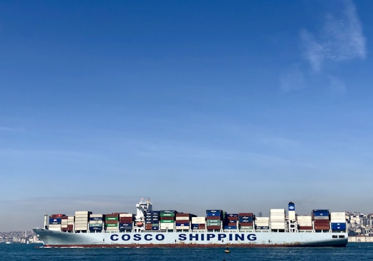 A large cargo ship with the name 'COSCO SHIPPING' is carrying numerous stacked shipping containers on calm blue waters. The background shows a distant coastline under a clear, blue sky.