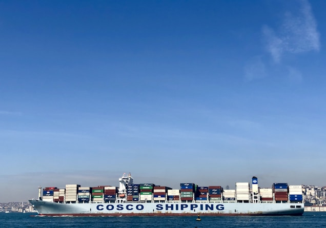 A large cargo ship with the name 'COSCO SHIPPING' is carrying numerous stacked shipping containers on calm blue waters. The background shows a distant coastline under a clear, blue sky.