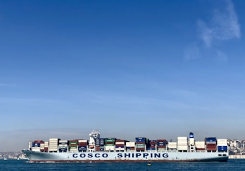 A large cargo ship with the name 'COSCO SHIPPING' is carrying numerous stacked shipping containers on calm blue waters. The background shows a distant coastline under a clear, blue sky.