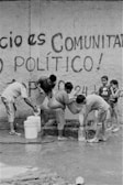 A group of people gather around a communal water source. Various individuals, including children, fill containers with water. The background features a wall with graffiti.
