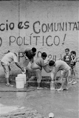 A group of people gather around a communal water source. Various individuals, including children, fill containers with water. The background features a wall with graffiti.