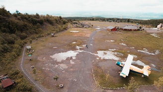 An aerial view of a grassy, open landscape featuring a yellow and white airplane parked on the ground. Surrounding the area are patches of grass, dirt paths, and puddles of water. On the right side, a few red-roofed buildings are visible, and on the left, there is a miniature train track running along the edge of the greenery.