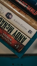A stack of hardcover books resting on a teal-colored surface. The visible titles include 'The Lost World' by T.G.H. James, a book by Adèle Geras, and 'Andy McNab' by Andrew Swanston. The spines display a range of colors from red to beige.