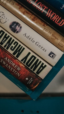 A stack of hardcover books resting on a teal-colored surface. The visible titles include 'The Lost World' by T.G.H. James, a book by Adèle Geras, and 'Andy McNab' by Andrew Swanston. The spines display a range of colors from red to beige.