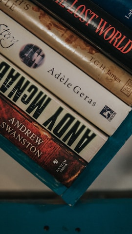 A stack of hardcover books resting on a teal-colored surface. The visible titles include 'The Lost World' by T.G.H. James, a book by Adèle Geras, and 'Andy McNab' by Andrew Swanston. The spines display a range of colors from red to beige.