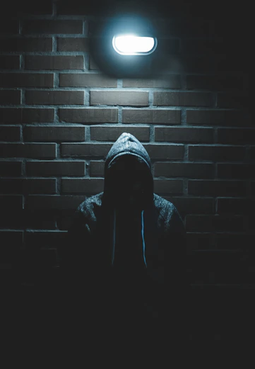 A person in a hooded sweatshirt in silhouette against a dark brick wall