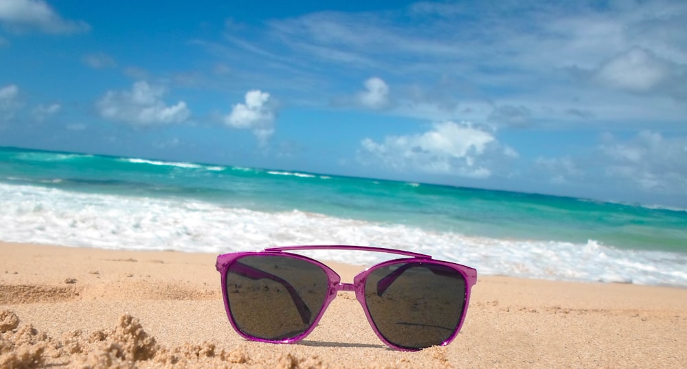 pink framed sunglasses on brown sand beside seashore during daytime