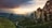 A stunning landscape features towering rock formations and a monastery perched on a cliff, with sunbeams breaking through a dramatic sky. Lush greenery and a winding path can be seen below, leading into a valley surrounded by mountains.