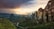 A stunning landscape features towering rock formations and a monastery perched on a cliff, with sunbeams breaking through a dramatic sky. Lush greenery and a winding path can be seen below, leading into a valley surrounded by mountains.