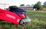 A vibrant red backpack with the brand name 'American Tourister' printed on it lies on a green grassy field. In the background, there is a multi-story building with a white facade and red accents, surrounded by trees under a clear blue sky.