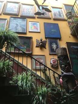 An indoor space features a staircase with black metal railings adorned with lush green plants. The walls are decorated with a variety of framed artworks including paintings and photographs. A wooden sculpture of a figure with butterfly wings is mounted on the wall. Multiple fish-shaped decorations hang from the ceiling, adding to the artistic ambiance. The wall colors are warm, primarily yellow, complementing the overall eclectic decor.