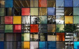 A grid of colorful, semi-transparent glass panels forms a wall, with each panel showing a variety of colors such as red, yellow, blue, and green. The light filtering through the glass creates patterns and the outlines of plants can be seen in the background. A hint of an indoor setting with tables and chairs is visible through the panels.