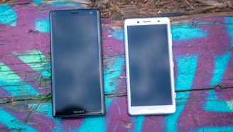 Two Sony smartphones are placed side by side on a vibrant, graffiti-covered wooden surface. The phone on the left is black, and the one on the right is white. The background includes bright colors such as purple, turquoise, and pink.