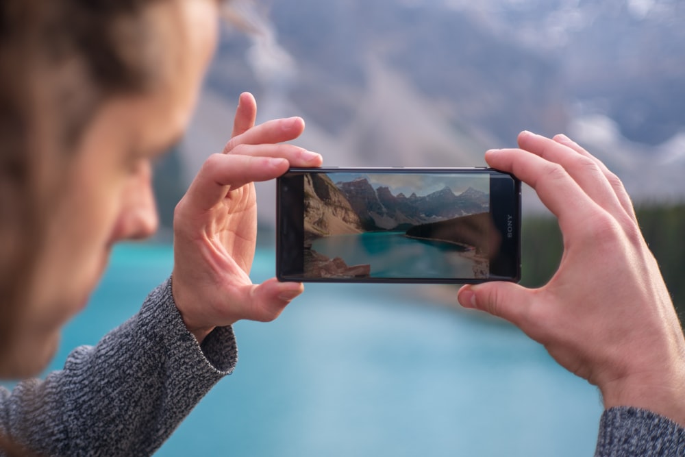 man capturing body of water and mountains from his phone