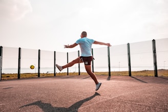 A person is energetically kicking a soccer ball mid-air on an outdoor court with wire fencing, under a clear sky. The background shows a scenic view of an open field and a body of water.