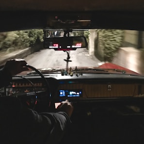 A view from inside a vehicle, showing the dashboard and steering wheel. The interior is dimly lit with visible dials and indicators glowing. A rearview mirror hangs above, reflecting part of the road behind. The driver’s hand is visible on the steering wheel. The road outside appears narrow and surrounded by greenery.