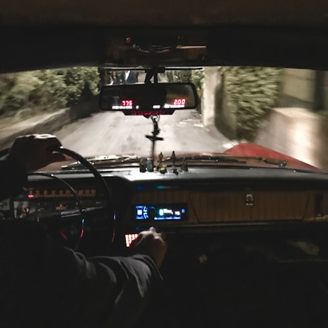 A view from inside a vehicle, showing the dashboard and steering wheel. The interior is dimly lit with visible dials and indicators glowing. A rearview mirror hangs above, reflecting part of the road behind. The driver’s hand is visible on the steering wheel. The road outside appears narrow and surrounded by greenery.