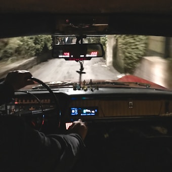 A view from inside a vehicle, showing the dashboard and steering wheel. The interior is dimly lit with visible dials and indicators glowing. A rearview mirror hangs above, reflecting part of the road behind. The driver&rsquo;s hand is visible on the steering wheel. The road outside appears narrow and surrounded by greenery.