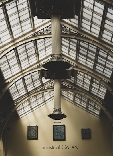 An interior view of a gallery with a high, arched ceiling made of glass panels. A large, decorative column extends from the ceiling down towards the floor. Several framed patterns are hung on the cream-colored walls, and the words 'Industrial Gallery' are visible below them.