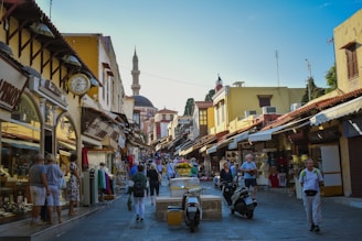 A bustling street scene in a historic town, featuring narrow cobblestone streets lined with shops selling souvenirs, clothes, and jewelry. People, including tourists, are walking around, shopping, and exploring. The architecture is a mix of stone and wooden buildings with a prominent mosque in the background, indicating a cultural or historical district.