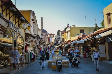 A bustling street scene in a historic town, featuring narrow cobblestone streets lined with shops selling souvenirs, clothes, and jewelry. People, including tourists, are walking around, shopping, and exploring. The architecture is a mix of stone and wooden buildings with a prominent mosque in the background, indicating a cultural or historical district.