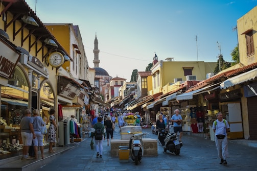 A bustling street scene in a historic town, featuring narrow cobblestone streets lined with shops selling souvenirs, clothes, and jewelry. People, including tourists, are walking around, shopping, and exploring. The architecture is a mix of stone and wooden buildings with a prominent mosque in the background, indicating a cultural or historical district.