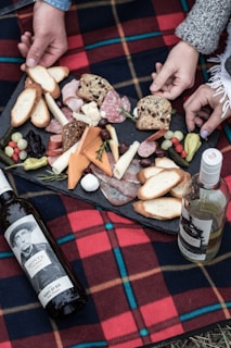 A picnic arrangement on a red and black checkered blanket features an assortment of cheeses, cold cuts, breads, and fruits on a slate platter. Several hands are reaching in, suggesting a shared dining experience. Two bottles of wine, one red and one white, are positioned next to the platter.