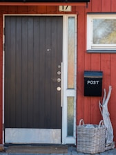 A wooden door with a dark finish and a metal strip at the bottom is centered in a red façade with vertical paneling. Beside the door, there is a black mailbox labeled 'POST' and a wicker basket containing some driftwood. A small rectangular window is positioned on the upper right side of the door, and a narrow pane of textured glass is on the left side of the mailbox.