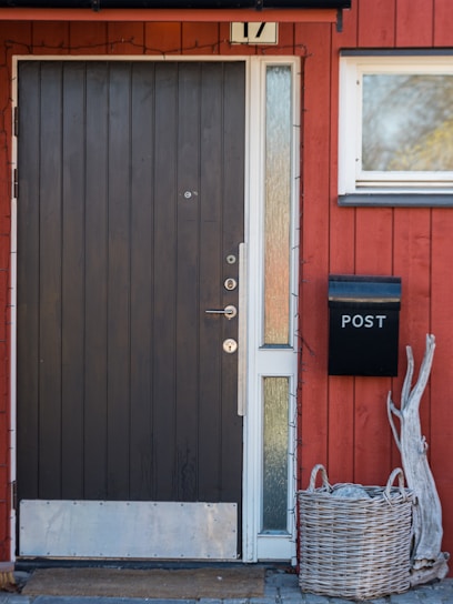 A wooden door with a dark finish and a metal strip at the bottom is centered in a red façade with vertical paneling. Beside the door, there is a black mailbox labeled 'POST' and a wicker basket containing some driftwood. A small rectangular window is positioned on the upper right side of the door, and a narrow pane of textured glass is on the left side of the mailbox.
